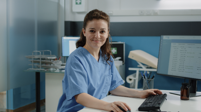 a white woman in light blue scrubs smiles at the camera while standing at a computer desk